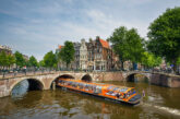 Busy canal cruise boat passing under a stone bridge on the Amsterdam Grachtengordel canal ring, with Golden Age merchant houses in the background