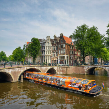 Busy canal cruise boat passing under a stone bridge on the Amsterdam Grachtengordel canal ring, with Golden Age merchant houses in the background