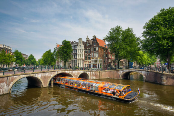 Busy canal cruise boat passing under a stone bridge on the Amsterdam Grachtengordel canal ring, with Golden Age merchant houses in the background