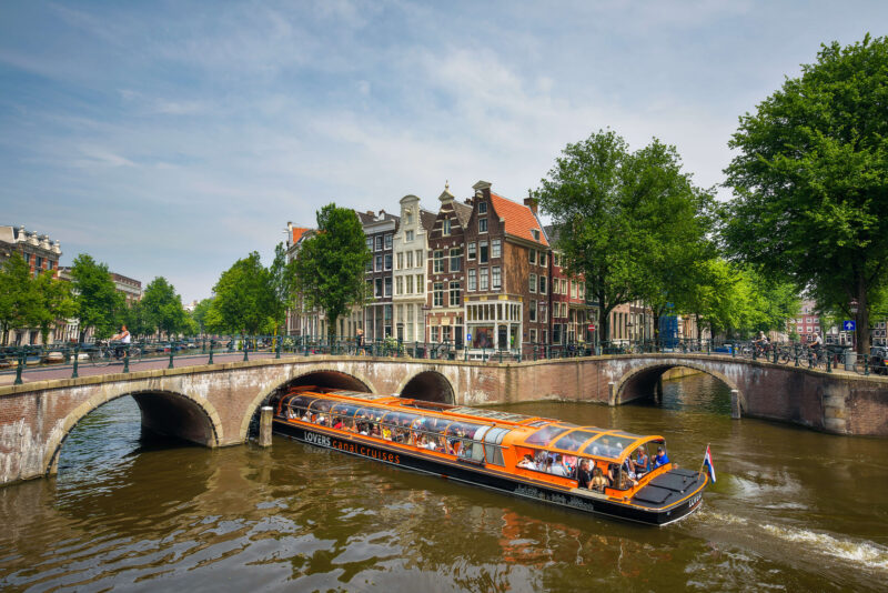Busy canal cruise boat passing under a stone bridge on the Amsterdam Grachtengordel canal ring, with Golden Age merchant houses in the background