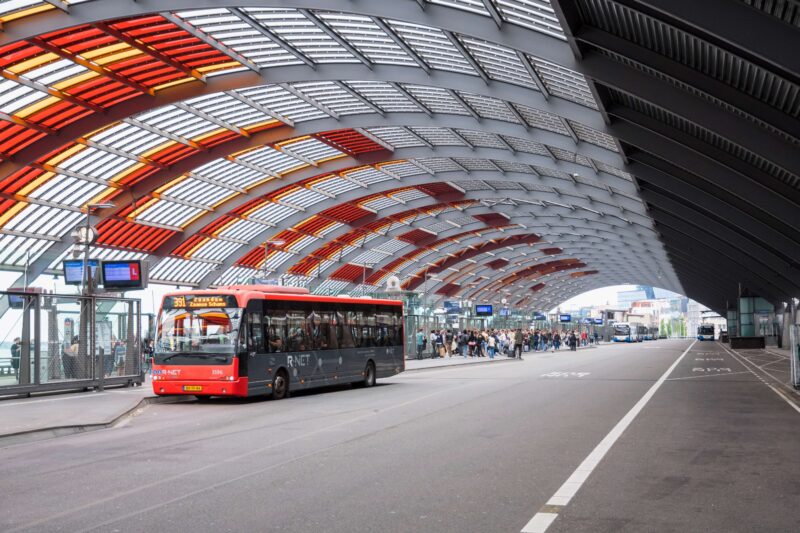 R-NET bus at Amsterdam Centraal station bus terminal under the curved architectural roof, connecting IJmuiden cruise port to Amsterdam city centre