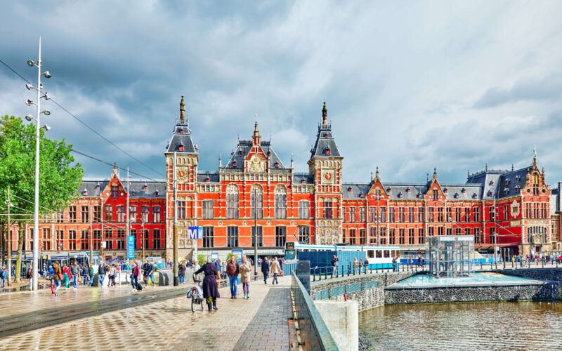 The iconic red-brick facade of Amsterdam Centraal station with trams, cyclists and visitors on the busy Stationsplein square