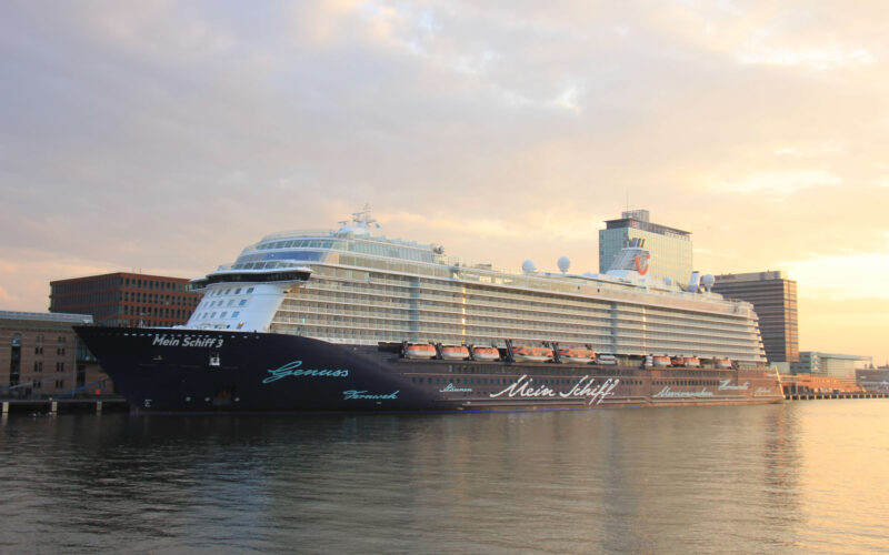 Mein Schiff 3 cruise ship docked at the Passenger Terminal Amsterdam (PTA) on the IJ waterfront at sunset, with the Amsterdam skyline in the background
