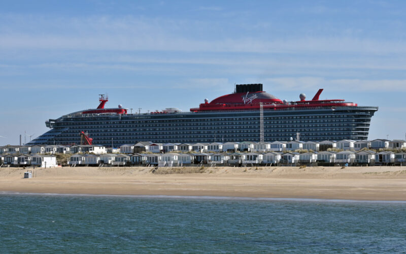 Virgin Voyages cruise ship towering over the beach cabins at IJmuiden aan Zee, with the North Sea in the foreground and the Felison Cruise Terminal behind