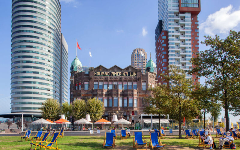 The historic Hotel New York building with Holland Amerika Lijn signage on Wilhelminapier, Rotterdam, surrounded by modern skyscrapers and deck chairs on the waterfront
