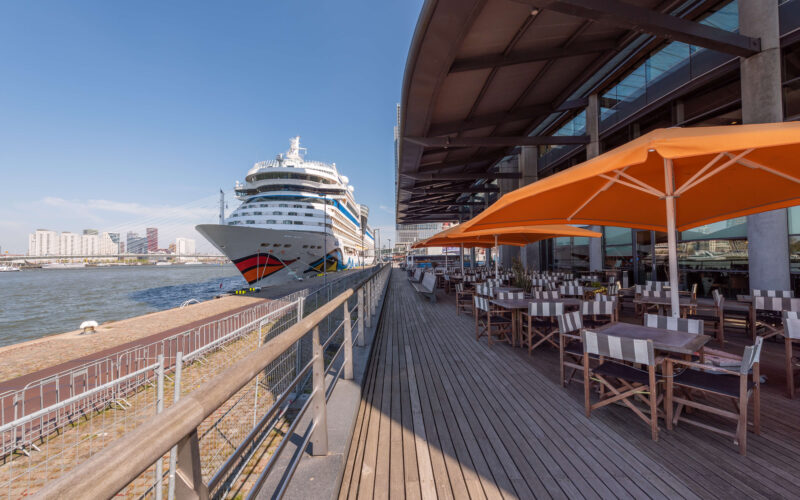Waterfront restaurant terrace with orange parasols on Wilhelminapier Rotterdam, with an AIDA cruise ship docked and the Erasmus Bridge in the background